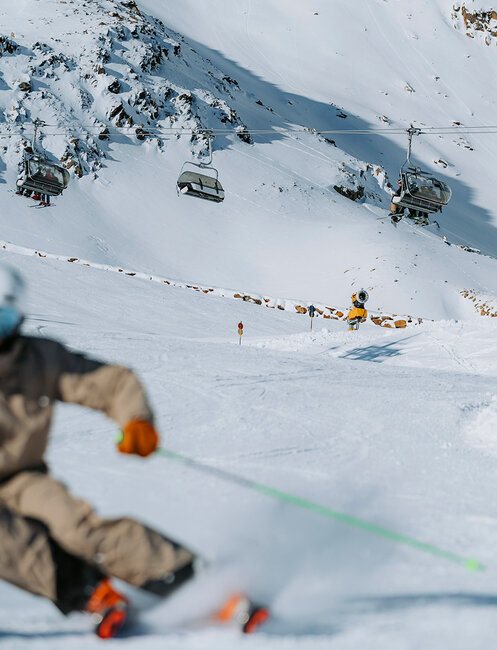 A skier in brown gear speeds down the slope, leaving a blur in the foreground. In the background, snow-capped mountains, ski lifts and other skiers are under a clear sky.