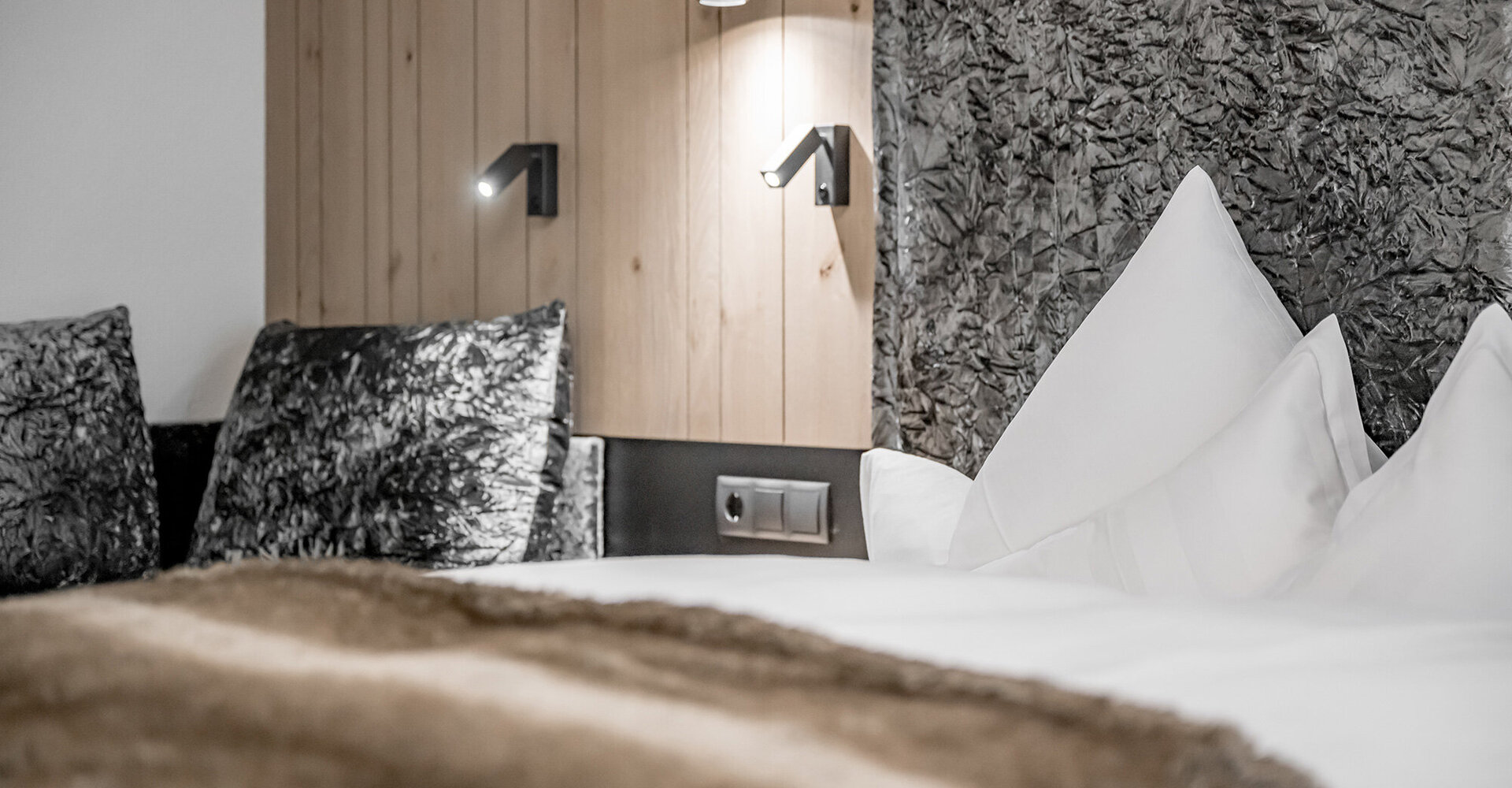 Close-up of a modern hotel bed with white pillows, a soft brown blanket, textured gray pillows, wood paneling, black wall lamps and a textured accent wall in the background.
