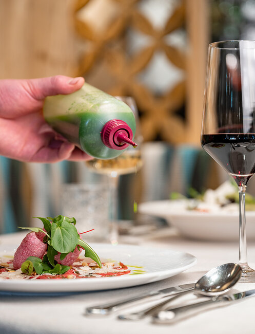 A hand pours green sauce from a squeeze bottle onto a plate of leafy greens, which szands next to a glass of red wine on a set dining table.