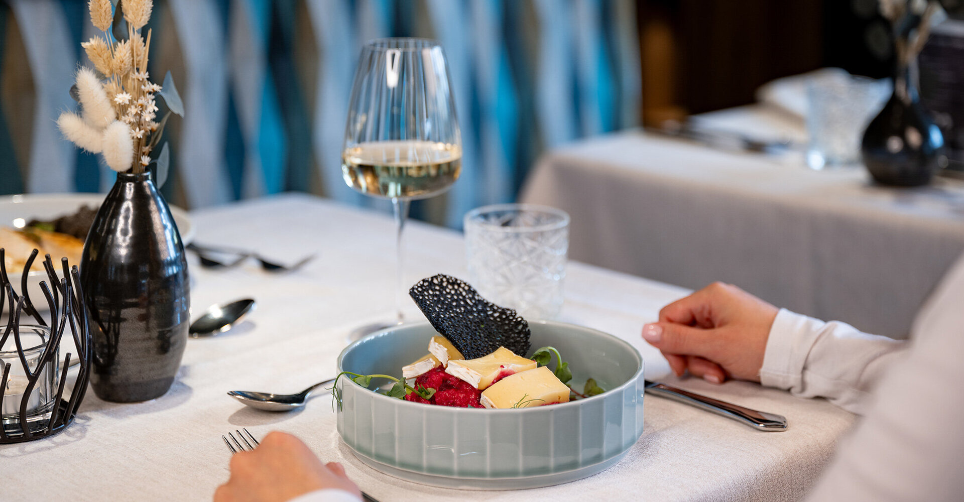 A person sits at a set dining table with a plate full of colorful delicacies, a glass of white wine and a vase of dried flowers.