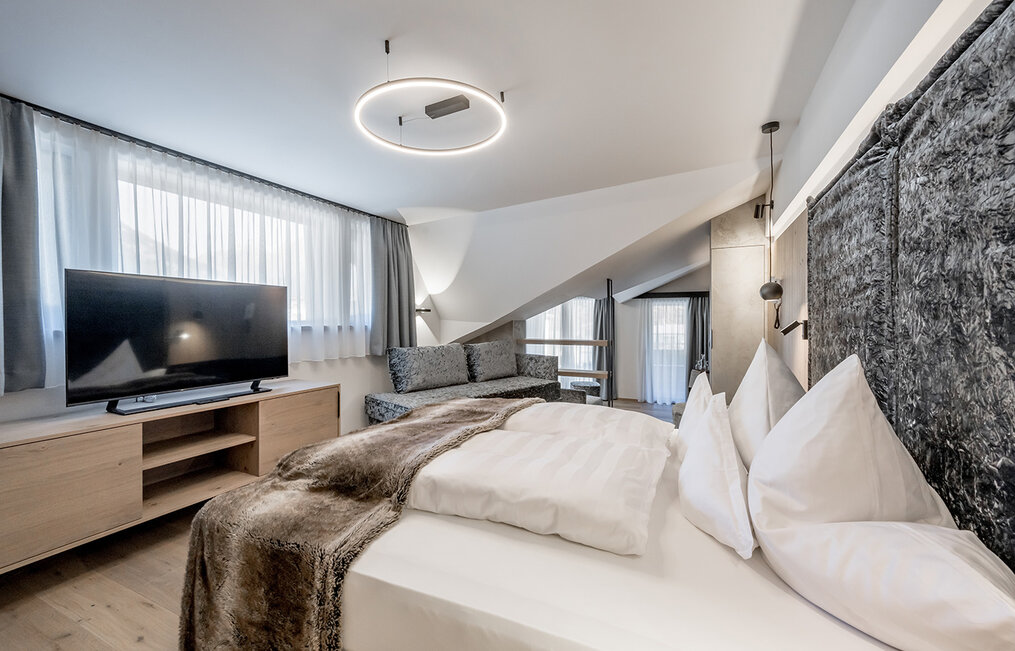 Bright hotel room featuring a large bed, modern lighting, a sofa, a TV on a wooden stand and large curtained windows, distinguished by contemporary décor and neutral colors.