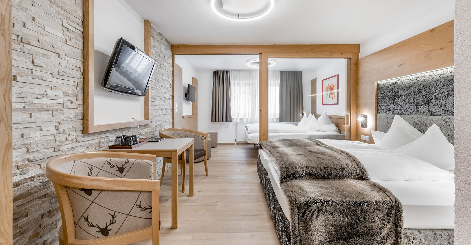 Modernes Hotelzimmer mit zwei Betten, einem Schreibtisch und Stühlen aus Holz, einem an der Wand montierten Fernseher und einer Akzentwand aus Stein. Große Fenster mit Vorhängen lassen Tageslicht herein. Die Einrichtung ist minimalistisch und in neutralen Farbtönen gehalten.