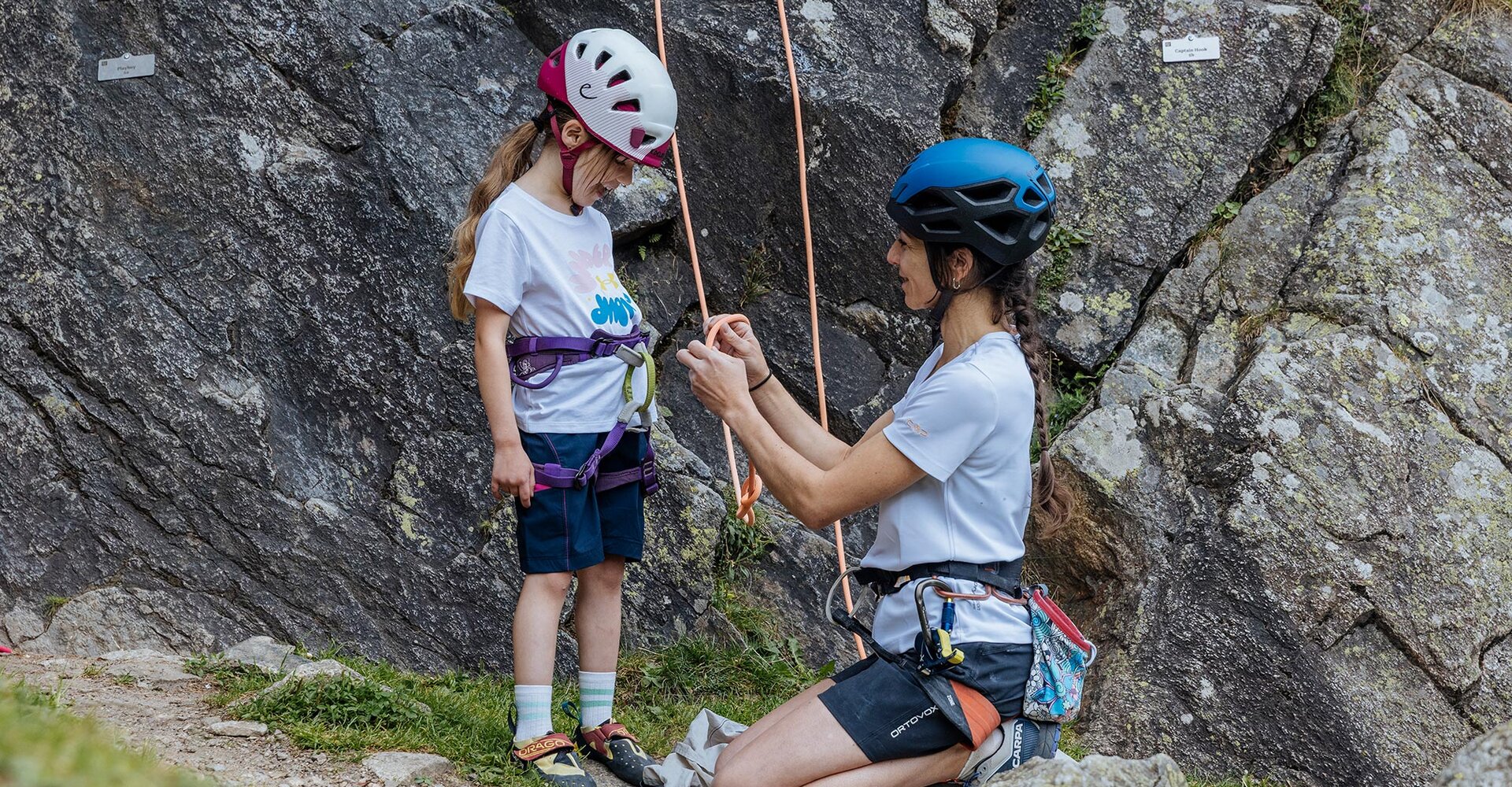 An adult is helping a child in climbing gear near a rock face. Both are wearing helmets and harnesses: the adult is kneeling and adjusting a rope while the child stands beside and watches.