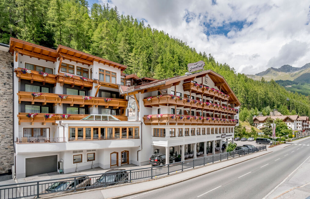A large hotel with wooden balconies and flower boxes is situated along a road, surrounded by green pines and mountains under a partly cloudy sky.