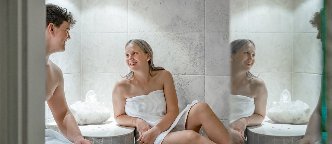 A man and a woman, dressed only in towels, are sitting in a steam sauna with tiled walls and talking. The woman smiles as she looks at the man.