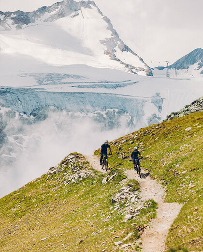 Two cyclists are riding along a narrow mountain path lined with green grass, while snow-capped peaks and a glacier are in the background under a cloudy sky.