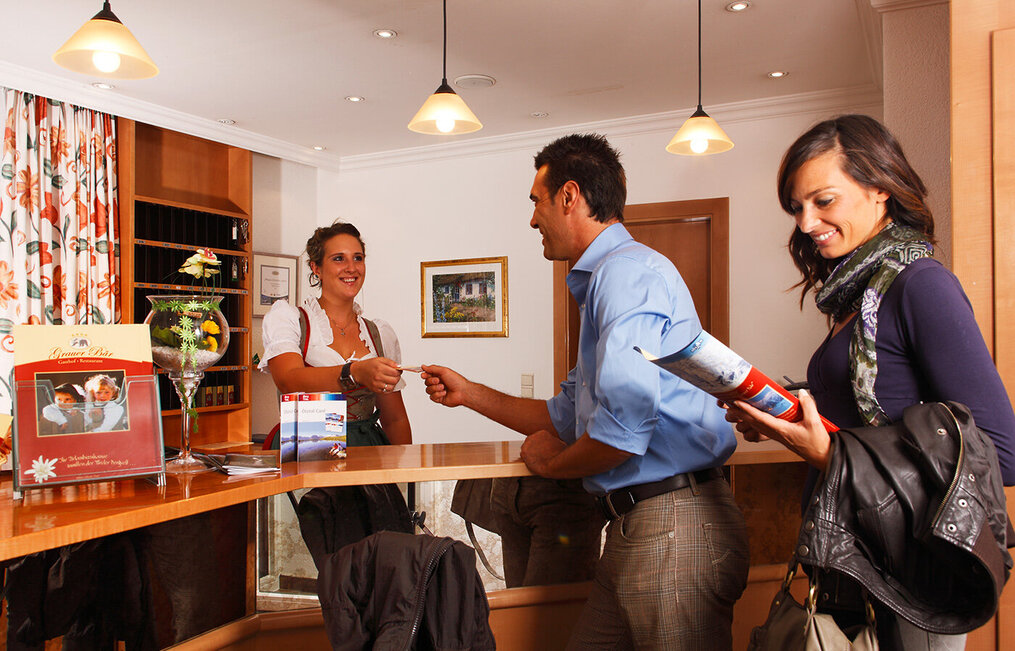 A man checks in at the hotel reception and hands something to the smiling receptionist, while a woman stands next to him with a magazine and her bag. Both are very happy.