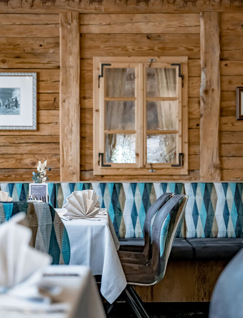 A cozy restaurant with wood-paneled walls, a window and tables set with white tablecloths and folded napkins. Modern chairs stand along the wall next to blue-patterned seating.
