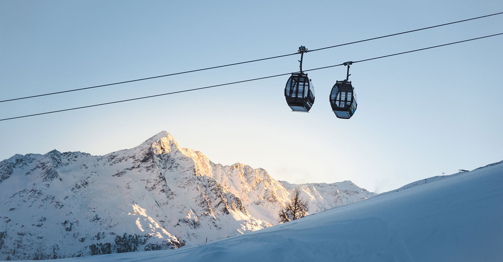 Two mountain gondola cabins float on a cable above a snowy mountain landscape, while in the background the sun illuminates the summit with a clear, light blue sky above them.