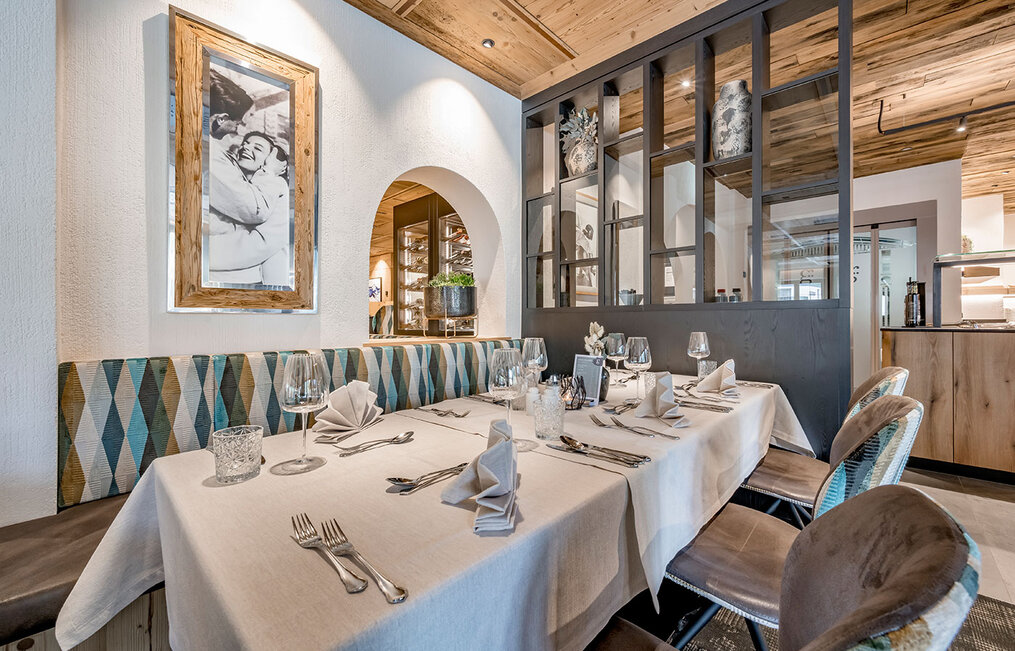 A cozy restaurant table for six, set with folded napkins, wine glasses and cutlery, featuring upholstered bench seating, wood accents and a framed black-and-white photograph on the wall.