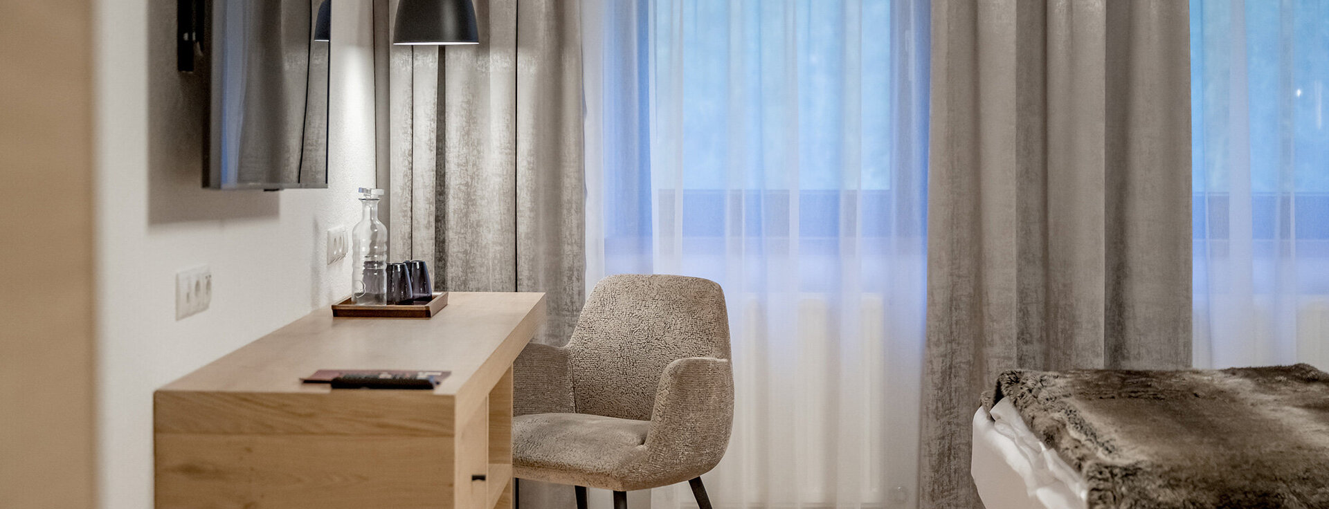 Ein modernes Hotelzimmer mit einem Schreibtisch, einem Polstersessel, einem an der Wand montierten Fernseher, einem Bett mit einer Felldecke sowie großen Fenstern mit transparenten Vorhängen und Vorhängen.