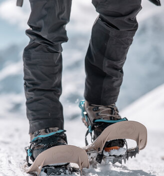 A person wearing black trousers and crampons attached to the boots, walking on snow. The background is snowy and mountainous.