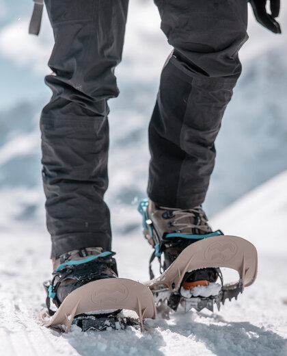 A person wearing black trousers and crampons attached to the boots, walking on snow. The background is snowy and mountainous.