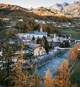 A mountain village with a river, a church and trees in autumn colors nestles in a green valley, surrounded by wood-covered hills and distant mountains under a brilliant blue sky.