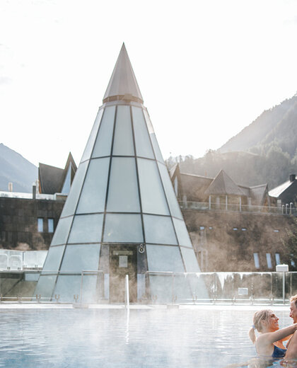 A couple embraces in a steaming outdoor pool while a pyramid-shaped glass building and mountains are in the background with a brilliant blue sky overhead.