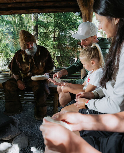 A group of people, including a man in a brown fur suit, are sitting together under a wooden shelter, shaping bread. Outdoors in a wooded setting, they are completely engrossed in their work.