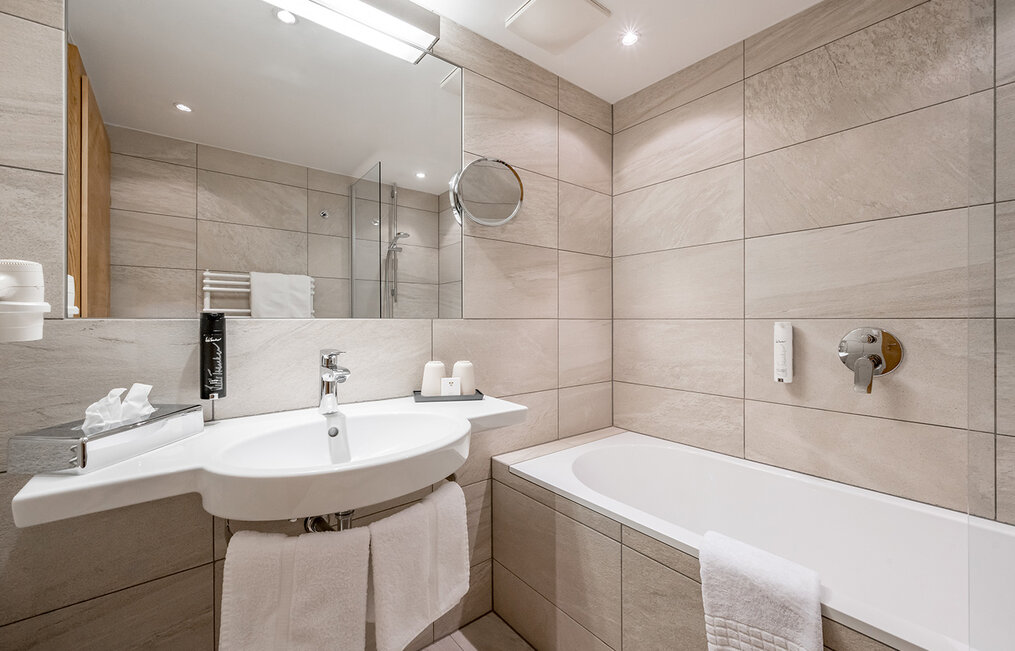 Modern bathroom with beige-tiled walls, a white sink with towels, a mirror, a bathtub and neatly arranged toiletries on the vanity.