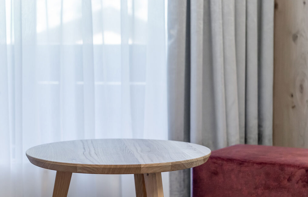 A round wooden table next to a red-upholstered stool, in front of delicate white curtains and light drapes in a softly lit room.