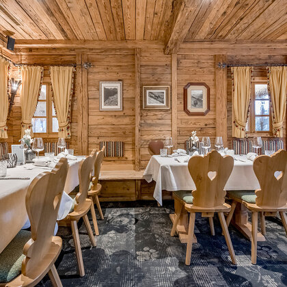 A cozy, rustic restaurant featuring wood-paneled walls, heart-shaped wooden chairs, white tablecloths and curtained windows. Framed pictures hang on the walls, the tables are set for dining.