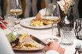 Eine Person sitzt in einem Restaurant beim Essen und hält Gabel und Messer über einem Teller mit Essen in der Hand. Auf dem Tisch stehen Weingläser, Wassergläser und eine Vase mit Trockenblumen; gegenüber ist ein weiterer Teller mit Essen zu sehen.