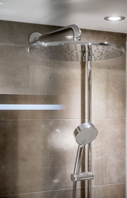 Modernes Duschsystem mit einem großen, runden Regen-Kopfbrausekopf und einer kleineren Handbrause, die beide an einer gefliesten Badezimmerwand unter einer Deckenleuchte angebracht sind.