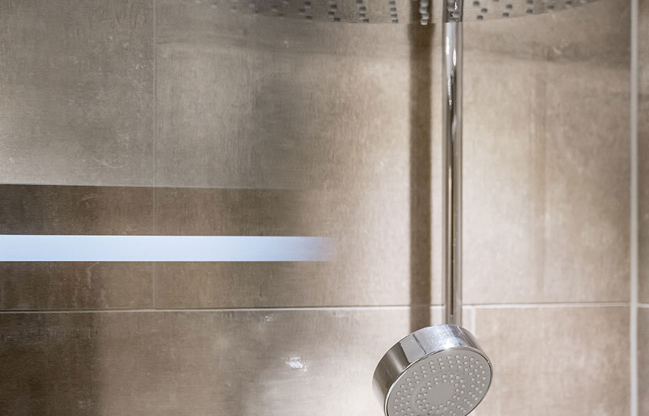 Modern shower system featuring a large, round rainfall showerhead and a smaller hand shower, both mounted on a tiled bathroom wall beneath a ceiling light.