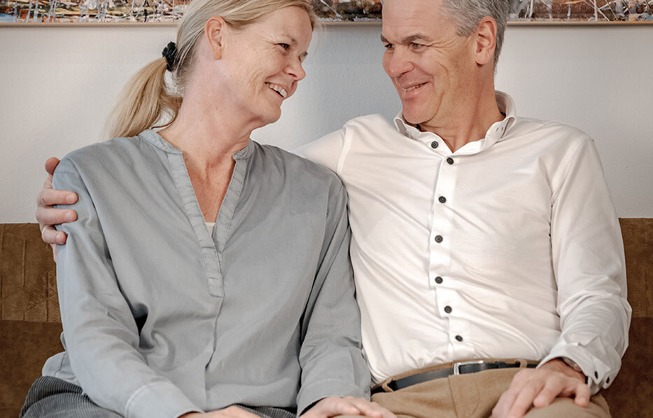 The host couple sits smiling close together on a sofa, looking at each other affectionately. The man has his arm around the woman. A painting of an animal hangs on the wall behind them.