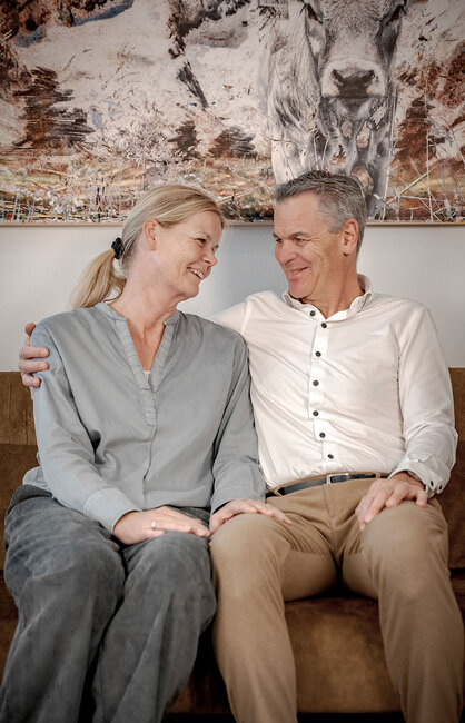 Das Gastgeber-Ehepaar sitzt lächelnd und eng beieinander auf einem Sofa und schaut sich liebevoll an. Der Mann hat seinen Arm um die Frau gelegt. An der Wand hinter ihnen hängt ein Tiergemälde.