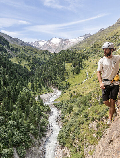 A person wearing a helmet and a climbing harness is climbing a rocky mountain face overlooking a green valley with a river flowing through it, surrounded by trees and distant snow-capped peaks.