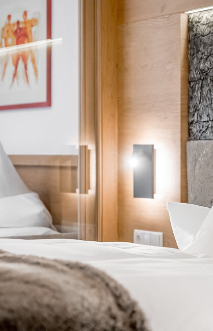 Ein modernes Hotelzimmer mit einem ordentlich gemachten Bett, weißen Kissen, einer flauschigen Decke, Holzakzenten, einem strukturierten Kopfteil und einem gerahmten Kunstwerk an der Wand. Sanfte Beleuchtung sorgt für eine gemütliche Atmosphäre.