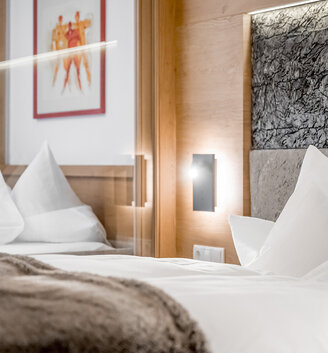 A modern hotel room featuring a neatly made bed, white pillows, a fluffy blanket, wood accents, a textured headboard and a framed piece of art on the wall. Soft lighting creates a cozy atmosphere.