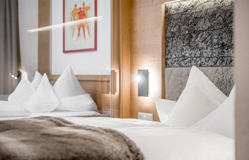 A modern hotel room featuring a neatly made bed, white pillows, a fluffy blanket, wood accents, a textured headboard and a framed piece of art on the wall. Soft lighting creates a cozy atmosphere.