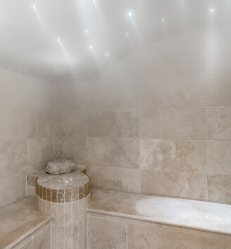 A steam room with tiled walls and benches, where gentle steam fills the space. Small lights are recessed into the ceiling, creating a relaxing atmosphere. A water fixture is mounted on the right-hand wall.