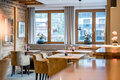 Gemütlicher Barbereich mit Holzwänden, beigen Stühlen und gepolsterten Bänken. Große Fenster mit Vorhängen lassen Tageslicht herein. Blumenvasen schmücken die Tische und Fensterbänke.