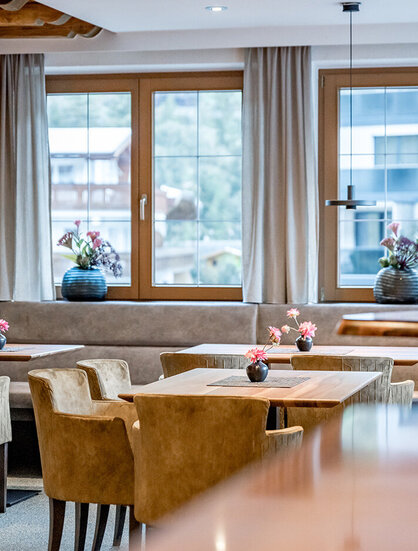 Cozy bar area with wood-paneled walls, beige chairs and upholstered benches. Large windows with curtains let in natural light. Flower vases adorn the tables and windowsills.