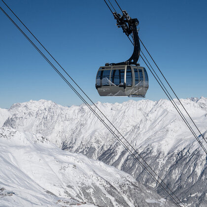A mountain gondola cabin travels over snow-capped mountain peaks beneath a brilliant blue sky, while rugged, snow-covered slopes and ridges stretch into the distance.