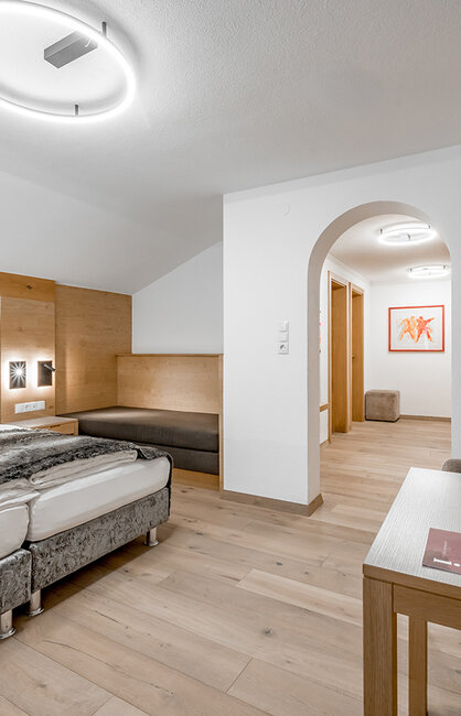 Modernes Hotelzimmer mit einem Doppelbett, einem grauen Kopfteil, einer Felldecke, einem kleinen Sofa, einem Schreibtisch mit Stuhl, einem Fernseher und Glasgeschirr. Helle Holzböden und Wände, mit einer gewölbten Türöffnung, die in einen Flur führt.