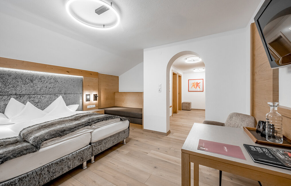 Modern hotel room featuring a double bed, a grey headboard, a faux-fur throw, a small sofa, a desk with a chair, a television and glassware. Light wood flooring and walls, an arched doorway leads into a hallway.