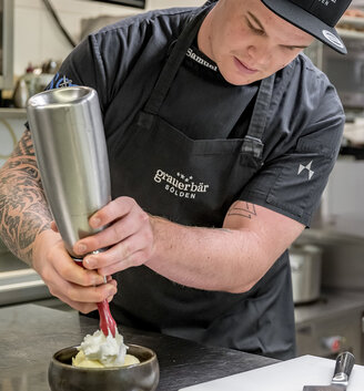A kitchen chef in a black apron and chef's hat squirts whipped cream into a bowl in a professional kitchen, concentrating fully on his work.