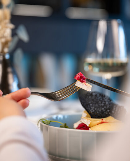 A person holds a fork and knife and cuts into a salad with cheese slices and leafy greens, at a set dining table featuring a wine glass and a decorative vase.