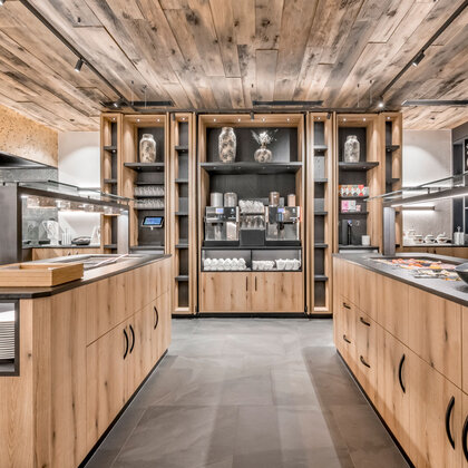 Modern buffet area featuring wooden cabinets, shelves with decorative vases and counters displaying coffee machines, plates, cups and breakfast essentials, all set beneath warm lighting and a natural wood ceiling.