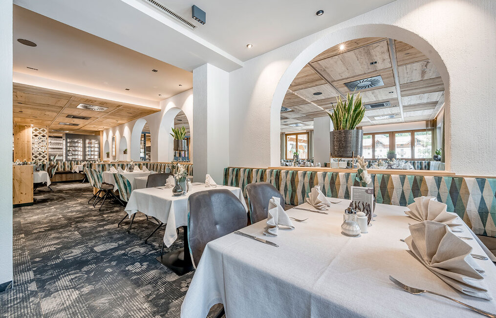 Modern restaurant interior featuring white tablecloths, neatly folded napkins, upholstered seating with geometric patterns, potted plants, large mirrors and arched wall openings. Warm lighting creates an inviting, elegant atmosphere.