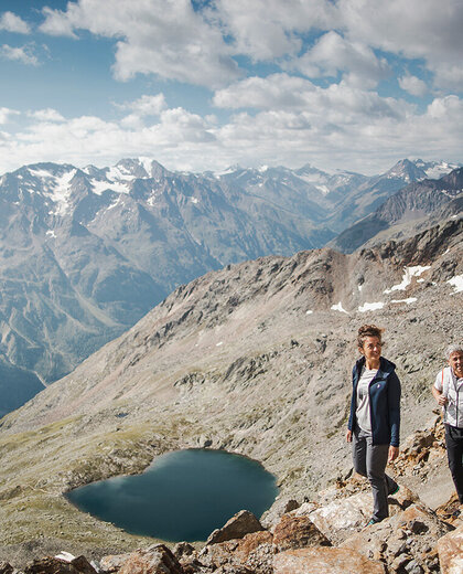 Two hikers are walking up a rocky mountain path with a heart-shaped lake and snow-capped peaks in the background, while the sky is partly cloudy.