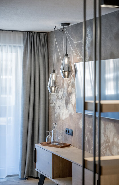 Eine moderne Hotelzimmer-Ecke mit einem Fernsehtisch aus Holz, zwei geometrischen Pendelleuchten, einem Fernseher vor einer Wand mit Blumenmuster und grauen Vorhängen neben einem Fenster mit weißen Gardinen.