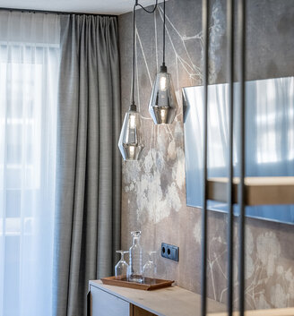 A modern hotel room corner featuring a wooden TV stand, two geometric pendant lights, a television set against a floral-patterned wall and gray drapes beside a window with white sheer curtains.