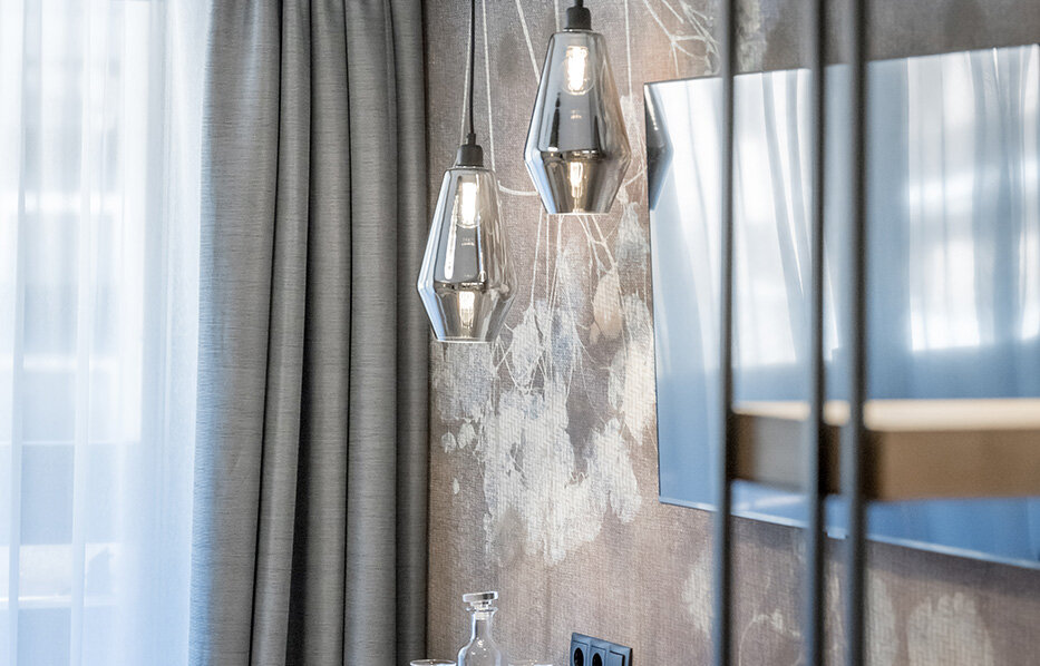 A modern hotel room corner featuring a wooden TV stand, two geometric pendant lights, a television set against a floral-patterned wall and gray drapes beside a window with white sheer curtains.