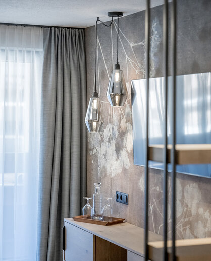 A modern hotel room corner featuring a wooden TV stand, two geometric pendant lights, a television set against a floral-patterned wall and gray drapes beside a window with white sheer curtains.