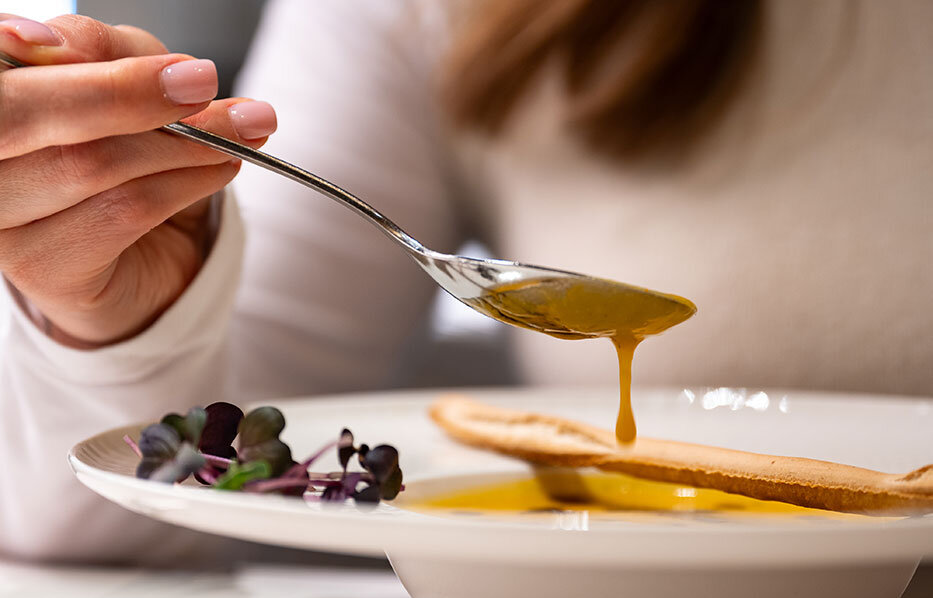 A person wearing a white turtleneck sweater lifts a spoonful of soup from a bowl. A crispy breadstick and microgreens lie on the plate. The soup is golden yellow and drips slightly from the spoon. 