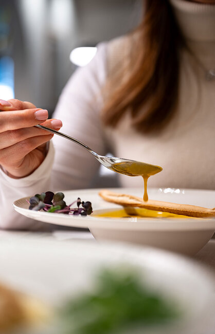 Eine Person in einem weißen Rollkragenpullover nimmt einen Löffel Suppe aus einer Schüssel, auf dem Teller liegen ein knuspriger Grissini und Microgreens. Die Suppe ist goldgelb, sie tropft leicht vom Löffel.