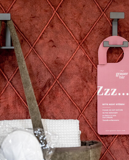 A pink door hanger and a bag containing towels hang on hooks in front of a red quilted wall.